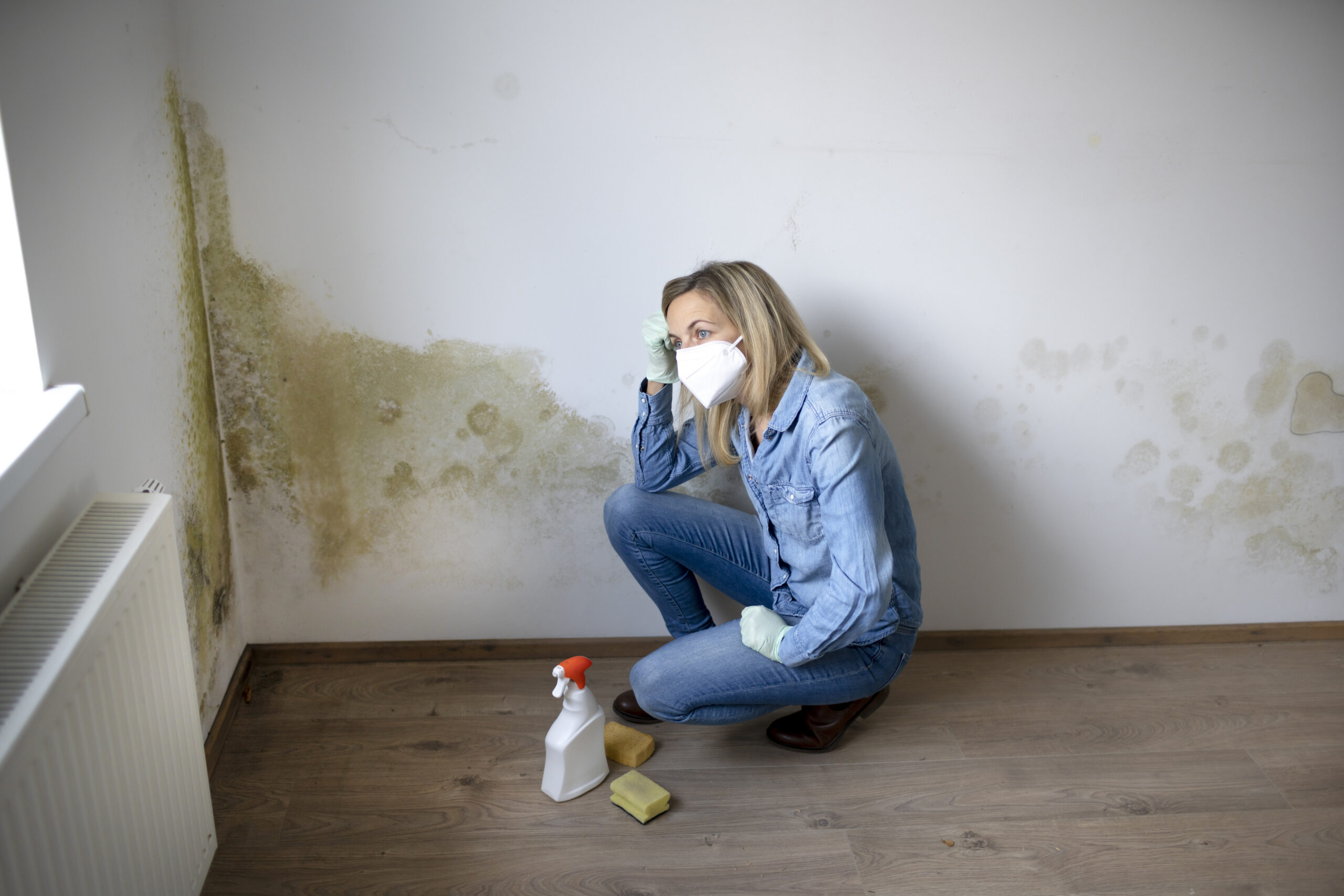 Blonde young woman is sitting in front of white apartment wall with mold on it and is frustrated